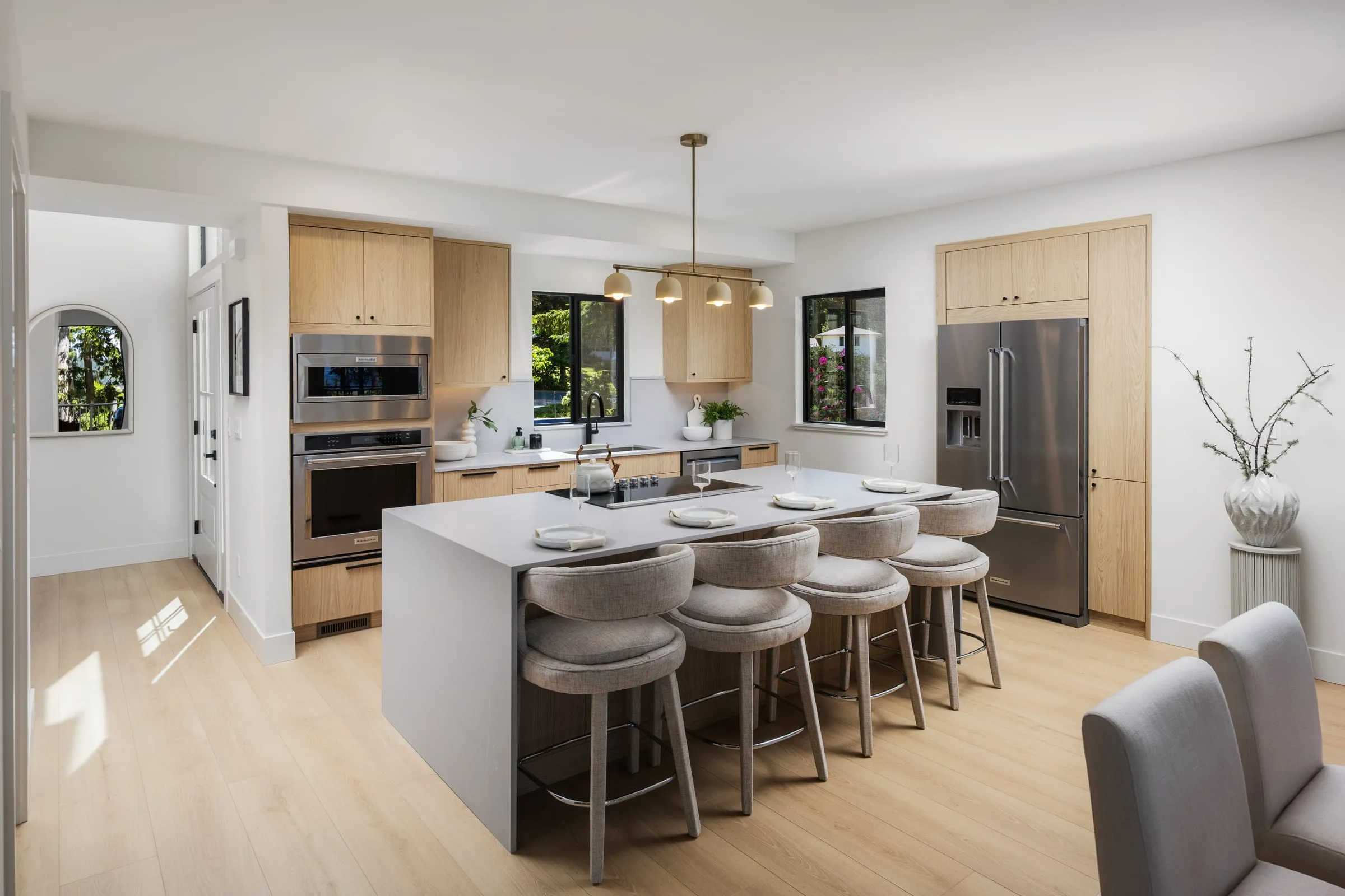 Pale oak kitchen with a long quartz island, six upholstered stools and stainless appliances