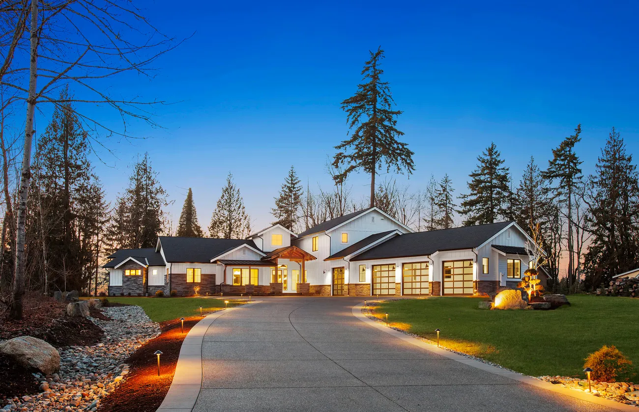 Single-storey Pacific Northwest home at twilight, lit driveway curving past evergreens to warm windows