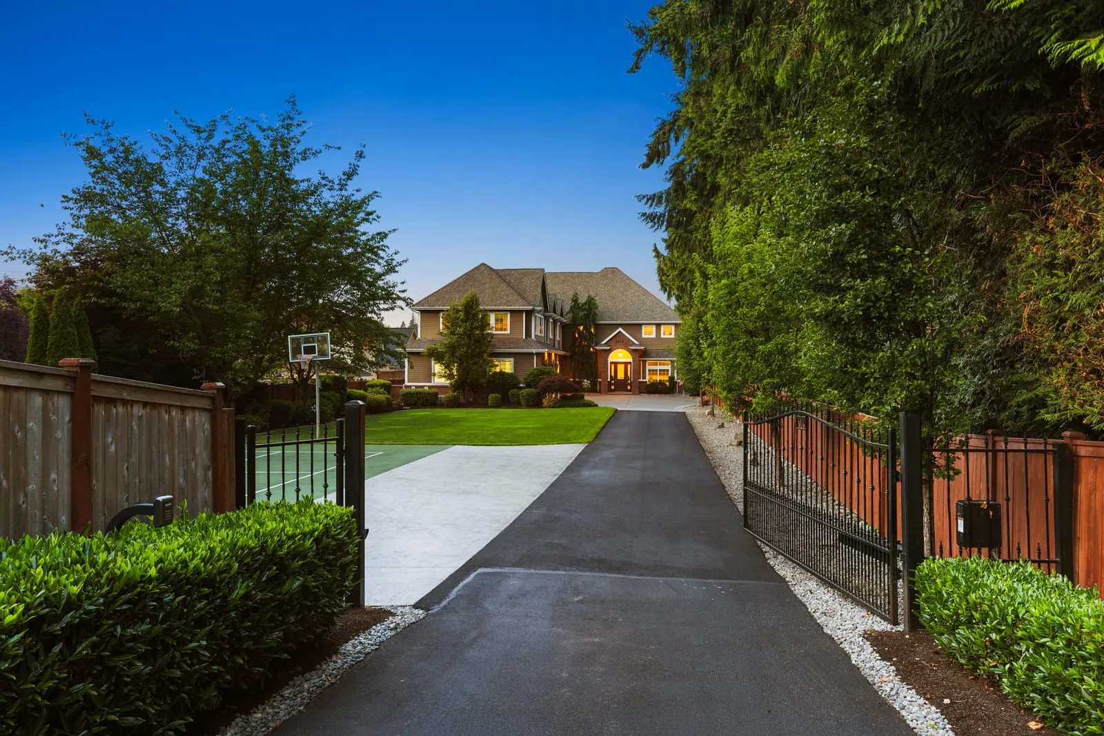 Long gated driveway through tall trees to a lit two-storey home at dusk