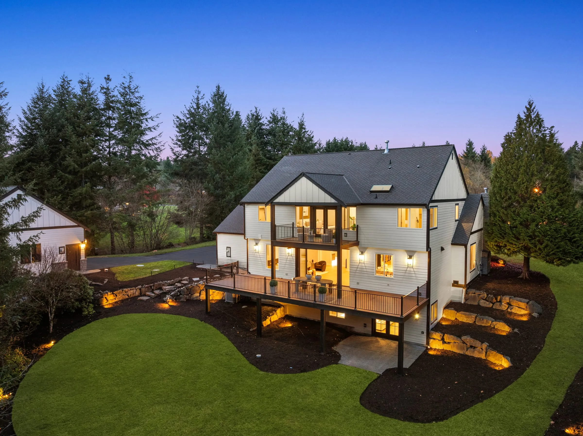 Aerial view at dusk of a white waterfront home with every window lit and a wraparound deck